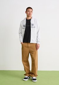 Grå zip-up hettegenser med logo, svart t-skjorte under, brune cordfløyelsbukser og svarte joggesko med hvite detaljer, stående på grønt gulv.