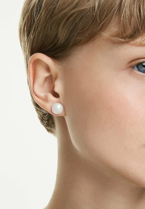 Nahaufnahme des rechten Ohrs einer Person, die einen einzelnen weißen Perlenstecker trägt, mit kurzen hellbraunen Haaren und teilweise sichtbarem blauem Auge.