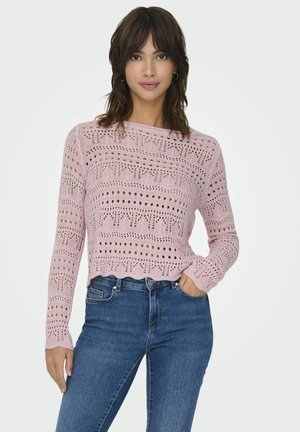 Maglione lavorato a maglia rosa chiaro con intricate fantasie traforate, maniche lunghe e orlo scallop, abbinato a jeans in denim blu.