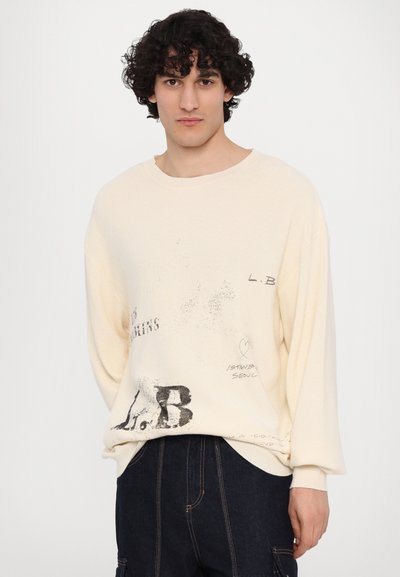 Pull en tricot couleur crème avec un col rond, orné de texte noir imprimé et de graphismes sur le devant. Coupe décontractée.
