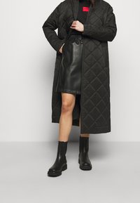 Schwarzer, gesteppter langer Mantel mit strukturiertem Finish, über einem schwarzen Leder-Minirock getragen und kombiniert mit knielangen schwarzen Stiefeln. Rotes Markenschild sichtbar.