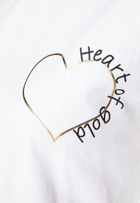 Camiseta de algodón blanca con un contorno de corazón metálico dorado y texto negro que dice "Corazón de Oro", con una textura suave y ligera.