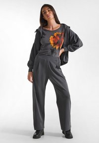 Graues Sportoutfit mit einem lässigen Oberteil mit einem orangefarbenen und gelben abstrakten Design und weitem Hosen, kombiniert mit schwarzen Sneakers.