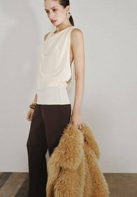 Top sin mangas de color crema con escote drapeado, combinado con pantalones anchos oscuros. La modelo lleva un abrigo de tono claro con textura similar a piel en las manos. Pulsera metálica llamativa.