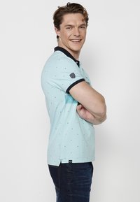 Svetlo modrá polo košeľa s tmavomodrým golierom, s vzorom malých trojuholníkov, krátkymi rukávmi a nášivkou loga na ľavom ramene.