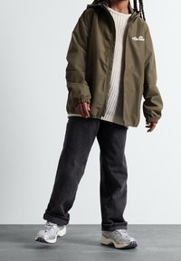 Osoba ubrana w oliwkową bluzę zapinaną na zamek, pod którą ma kremowy sweter dzianinowy, czarne luźne dżinsy oraz srebrno-białe sneakersy, stoi na szarym podłodze.