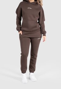 Set di felpa e pantaloni della tuta marroni in tessuto morbido, con una tasca frontale e testo del logo. I pantaloni hanno polsini elastici e una vestibilità rilassata.