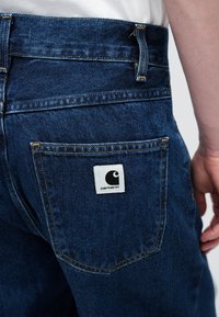 Jeans de denim en azul oscuro con una textura suave. Presentan una etiqueta blanca con un logo negro en el bolsillo trasero y costuras amarillas.