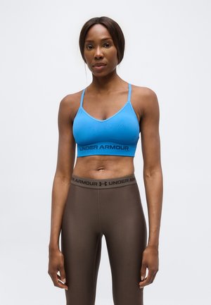 Kobieta w niebieskim sportowym biustonoszu Under Armour i brązowych legginsach z wysokim stanem z paskiem Under Armour, stojąca na jednolitym białym tle.