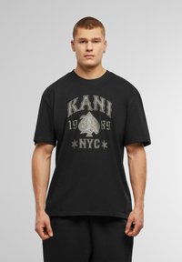 Svart bomullst-shirt med grafiskt tryck med "KANI," ett spelkortssymbol, och "NYC" i kontrasterande färger. Korta ärmar, avslappnad passform.
