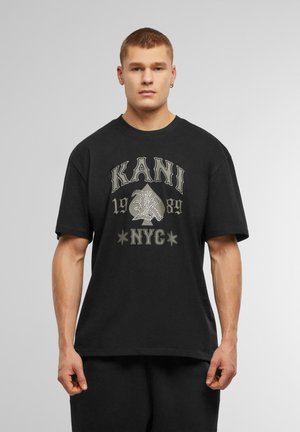 Zwarte katoenen T-shirt met een grafische print van "KANI," een speelkaart symbool en "NYC" in contrasterende kleuren. Korte mouwen, relaxed fit.