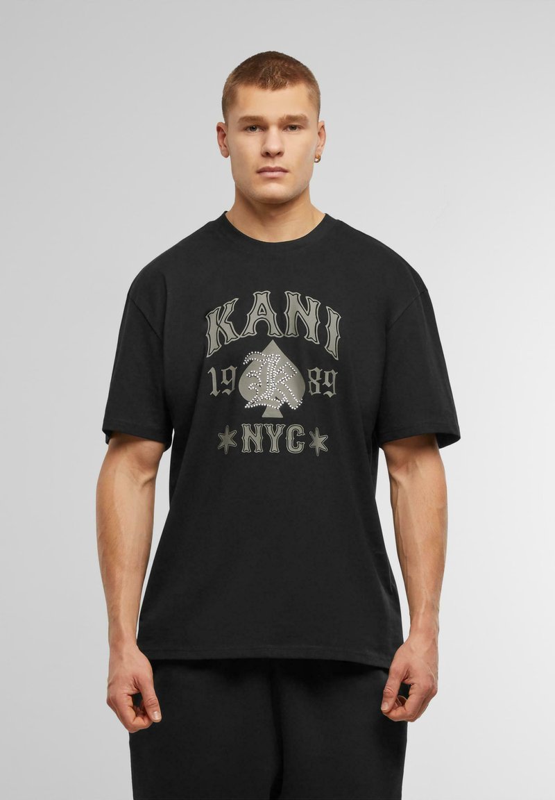 Svart bomullst-shirt med grafiskt tryck med "KANI," ett spelkortssymbol, och "NYC" i kontrasterande färger. Korta ärmar, avslappnad passform.