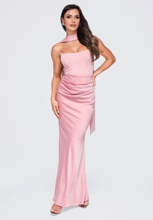 Vrouw met lang donker haar die een mouwloze roze satijnen avondjurk draagt met een gedrapeerde taille en een bijpassende halsdoek, staand met één hand op haar heup.
