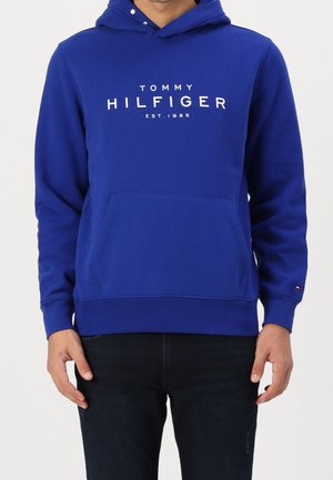 Person trägt einen blauen Tommy Hilfiger Hoodie mit Fronttasche und weißem Logo-Schriftzug, kombiniert mit schwarzer Hose, stehend vor schlichtem Hintergrund.