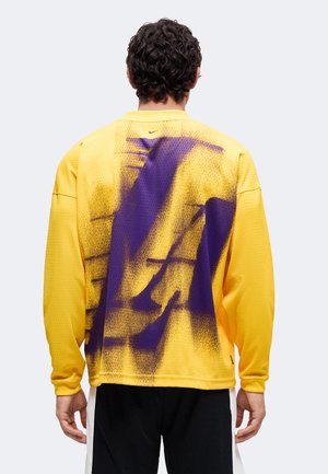 Homme aux cheveux bouclés portant un maillot de sport jaune à manches longues avec un grand motif abstrait violet dans le dos et un short noir.