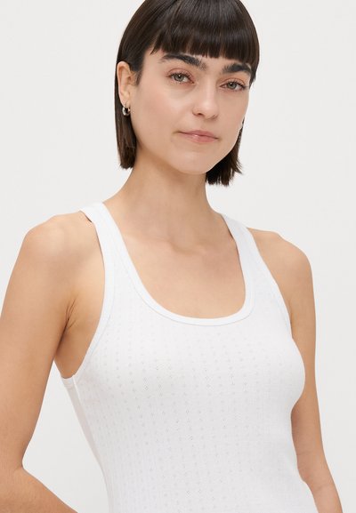 Balts ribots topiņš ar plānām siksniņām un izgriezumu ap kaklu. Satur mazus, vienmērīgi izkārtotus caurumus tekstūras un gaisa caurlaidības nodrošināšanai.