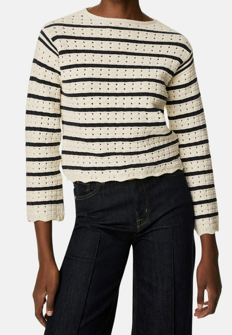 Sweter w paski kremowo-czarny z dzianiny z oczkowym wzorem, zestawiony z ciemnymi dżinsami z wysokim stanem na stojącej osobie.