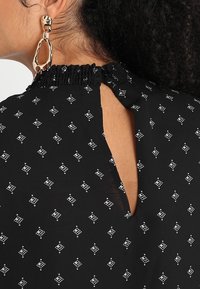 Blouse noire avec un col froncé et une petite fermeture à bouton à l'arrière, ornée d'un motif géométrique blanc.