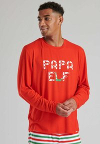 Camisa de manga larga roja con la inscripción "PAPA ELF" en letras inspiradas en bastones de caramelo, con acentos en verde. Tejido suave, corte cómodo.