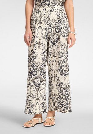 Wijde crèmekleurige broek met ingewikkelde zwarte bloemen- en ornamentpatronen, gedragen met lichte sandalen met bandjes en een zilveren armband aan de rechterpols.