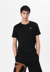 Black cotton crew neck t-shirt featuring a small red logo on the left chest. Short sleeves and a relaxed fit. Simple and minimalistic design.