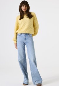 Vrouw met lang bruin haar, gekleed in een gestructureerd geel trui en lichtblauwe hoge taille wijde broek, staand tegen een witte achtergrond.