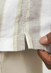 Camisa ligera de tejido a rayas en beige y blanco, con un dobladillo dividido y costura visible, y una mano sosteniendo el dobladillo.