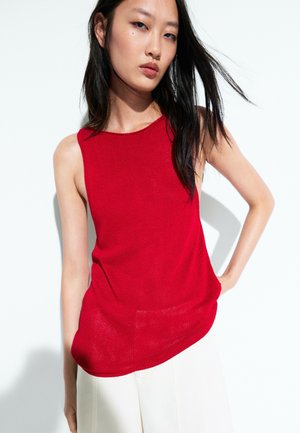 Donna dai lunghi capelli scuri indossa un top rosso senza maniche e pantaloni bianchi, in piedi contro uno sfondo chiaro e uniforme, guardando avanti.