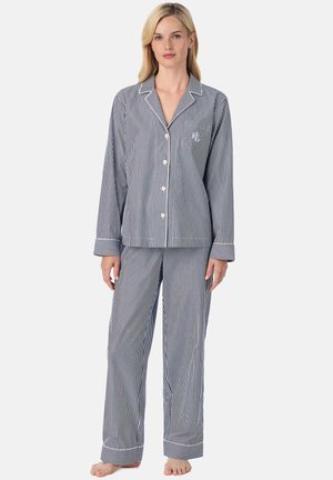 Vrouw draagt een pyjama met verticale marineblauwe en witte strepen, bestaande uit een bovenstuk met knopen en een lange broek, staat op blote voeten tegen een neutrale achtergrond.