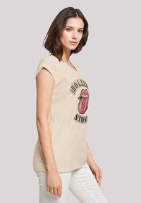 Beiges Baumwoll-T-Shirt mit rundem Halsausschnitt, kurzen Ärmeln, mit einem roten Lippen-Logo und schwarzem "ROLLING STONES"-Schriftzug.