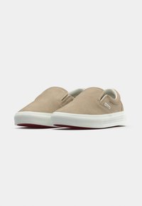 Paire de baskets beige en daim à enfiler avec semelles blanches en caoutchouc et dessous rouges sur un fond clair uni.