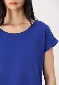 Nainen pukeutuneena yksiväriseen kuninkaansiniseen lyhythihaiseen t-paitaan, jossa on pyöreä pääntie, valkoista taustaa vasten.