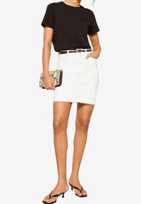 Žena v černém topu s krátkým rukávem, bílých roztrhaných denimových sukni s černým opaskem, černých sandálech na podpatku, držící kabelku z hadí kůže, pózující na bílém pozadí.