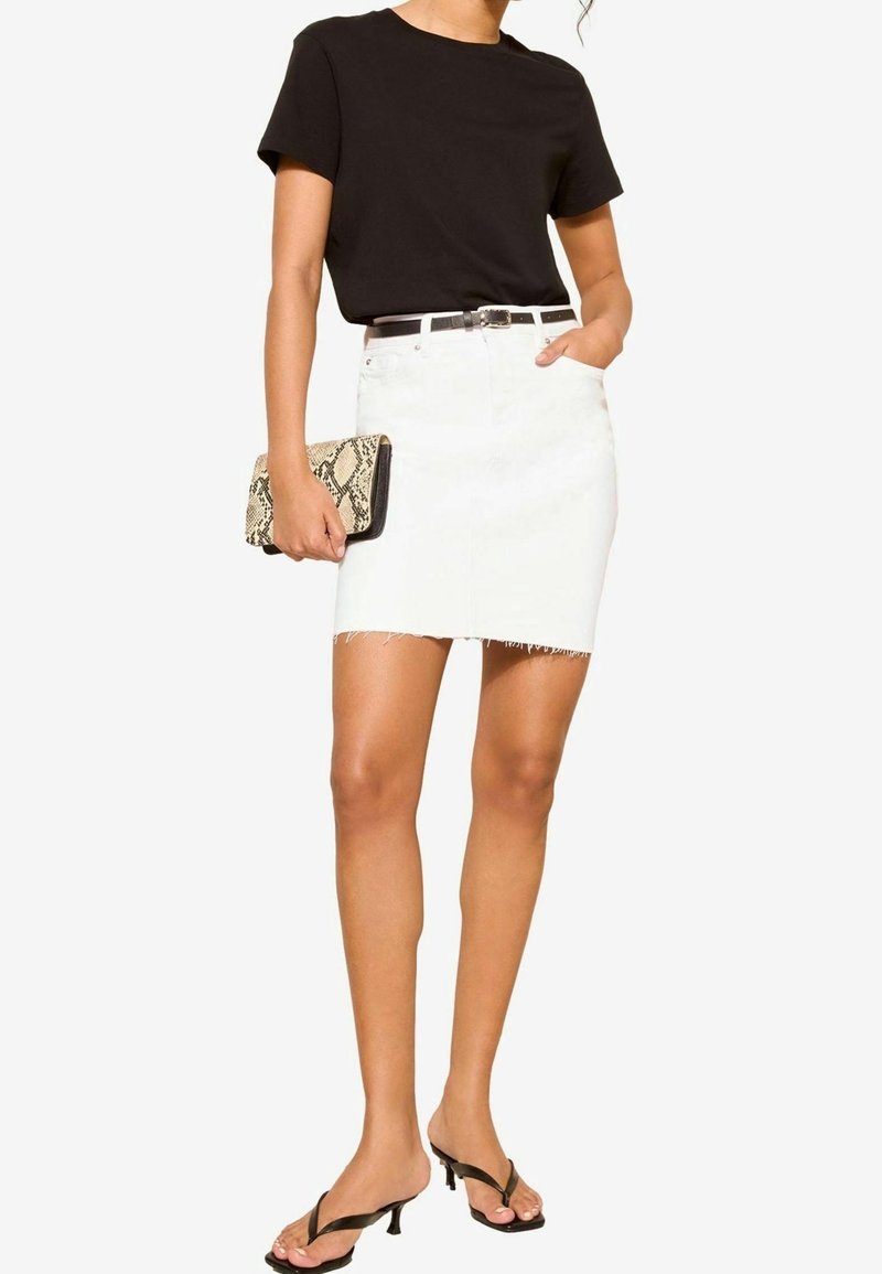 Žena v černém topu s krátkým rukávem, bílých roztrhaných denimových sukni s černým opaskem, černých sandálech na podpatku, držící kabelku z hadí kůže, pózující na bílém pozadí.