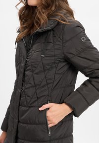 Schwarze, gesteppte Puffjacke mit einem Reißverschluss vorne, elastischen Bündchen und verstellbaren Zugkordeln an der Kapuze. Inklusive Seitentaschen.