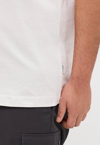 Camiseta de algodón blanca con dobladillo recto y etiqueta en la costura lateral, combinada con pantalones gris oscuro que cuentan con un bolsillo lateral.