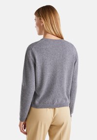 Šedý pletený svetr s kulatým výstřihem a dlouhými rukávy, který má měkkou texturu a volný střih, doplněný o světle zbarvené kalhoty.