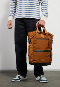 Mochila de nylon castanha com tecido texturizado, alças de transporte duplas, bolso frontal com fecho e acabamentos pretos na base. Usada com jeans escuros.