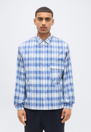 Homme portant une chemise à carreaux bleue et blanche à boutons, manches longues et une poche poitrine, debout devant un fond uni.
