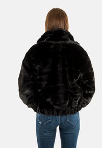 Veste en fausse fourrure noire avec un col montant et un ourlet élastique. Présente une texture douce et pelucheuse ainsi qu'une coupe décontractée. Portée avec un jean bleu.
