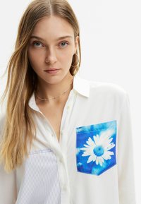 Camicia bianca con colletto a bottoni, caratterizzata da un design floreale blu sulla tasca. Il lato sinistro presenta un motivo a righe blu. Tessuto liscio, design casual.