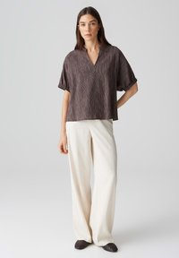Blusa di un marrone scuro testurizzata con scollo a V e maniche corte, abbinata a pantaloni bianchi a gamba larga e mocassini scuri a punta. Design semplice.