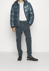 Chaqueta de plumas en color azul verdoso con acabado brillante, combinada con pantalones verdes oscuros y zapatillas negras con toques en rojo, blanco y azul. Camiseta blanca por debajo.