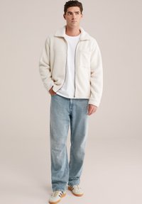 Jachetă albă din fleece cu fermoar, guler mare, buzunar frontal, combinată cu blugi de culoare albastră deschisă și teniși crem cu dungi negre. Fundal deschis.