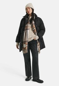 Schwarze Puffy-Jacke mit hohem Kragen, beigefarbener Strickpullover, gemusterter Karoschal und weit geschnittene schwarze Jeans. Schwarze Ankle Boots runden den Look ab.