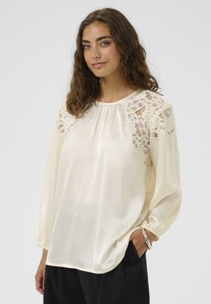 Žena u krem bluzi s čipkastim detaljima na ramenima i dugim rukavima, nosi crne hlače, stoji s jednom rukom u džepu.