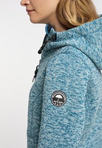 Blå texturerad sweatshirt med huva, med ett svart runt logotypmärke på ärmen och justerbara dragsko för huvan.