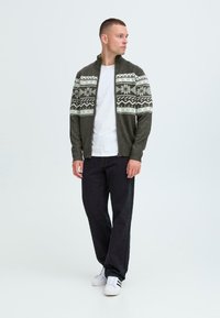 Stickad zip-up kofta i mörkgrön, med ett mönstrat fair isle-mönster i vitt. Kombinerad med svarta byxor och vita sneakers.