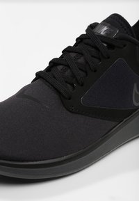 Zapatilla deportiva negra con parte superior de tela texturizada, acentos sintéticos y suela de goma. Cordones y logotipo en negro.