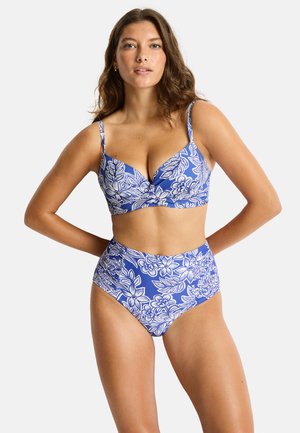 Frau trägt ein blau-weiß geblümtes Bikini-Set mit hohem Bund und steht mit den Händen hinter dem Rücken vor einem schlichten Hintergrund.